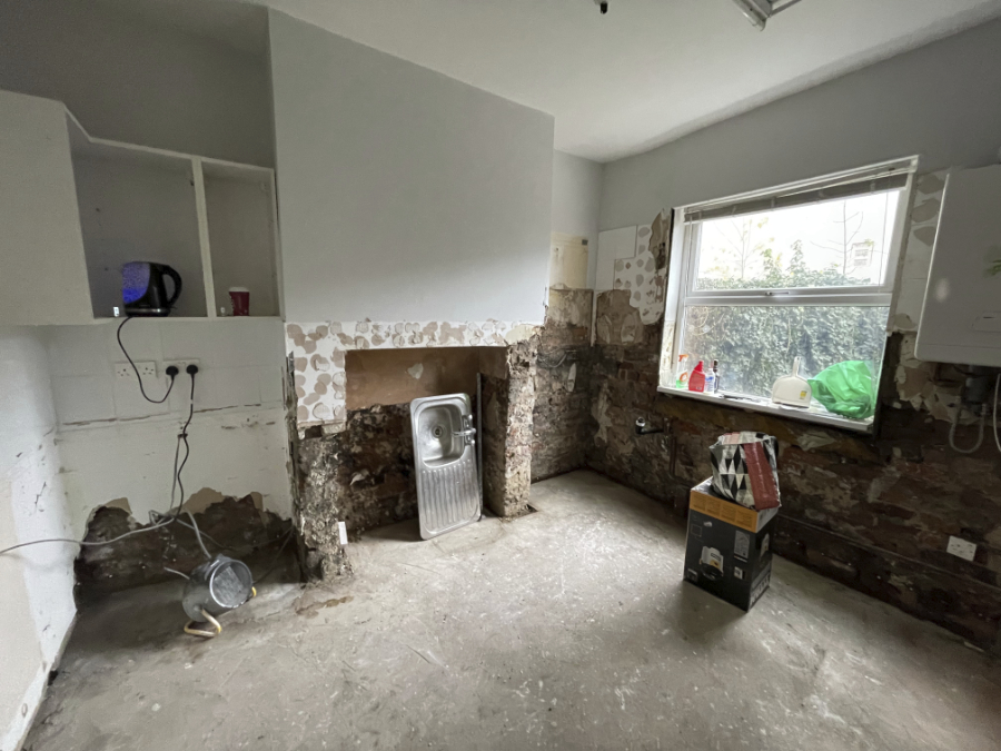 A 'before' shot of a kitchen renovation: bare concrete floor, exposed brick walls, visible wires and pipes, crumbling plaster